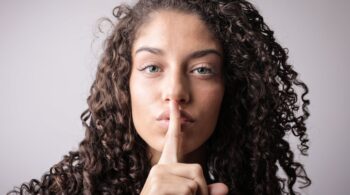 portrait photo of woman with brown curly hair doing the shhh sign