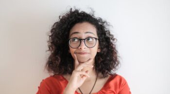 portrait photo of woman in red top wearing black framed eyeglasses standing in front of white background thinking