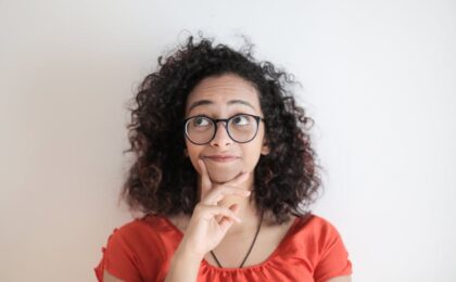 portrait photo of woman in red top wearing black framed eyeglasses standing in front of white background thinking