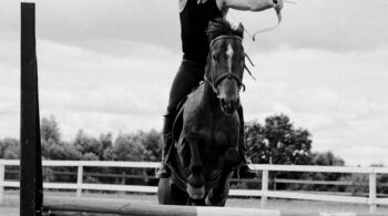 grayscale photo of man shooting an arrow while riding a horse